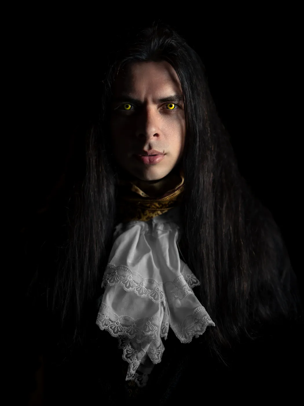 A long-haired person in a dark coat and white lace cravat, with yellow theatrical eyes.