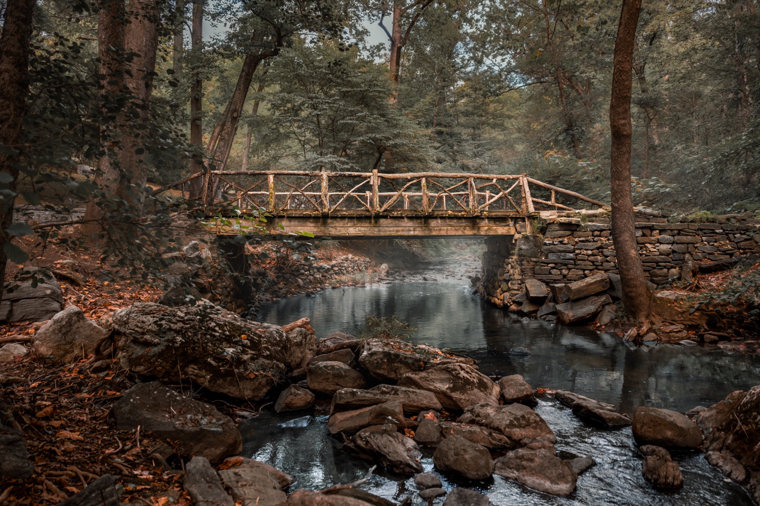 A rustic wooden bridge over a rocky stream in autumn woodland.